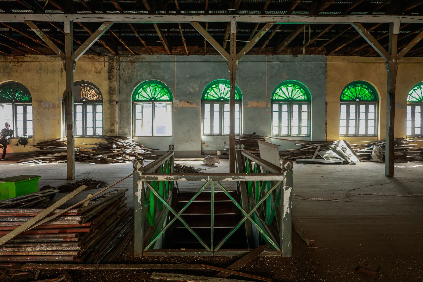 The main hall of YWCA Yangon during restoration, with steel roof trusses, green arched fanlight windows and timber stacked on the floor