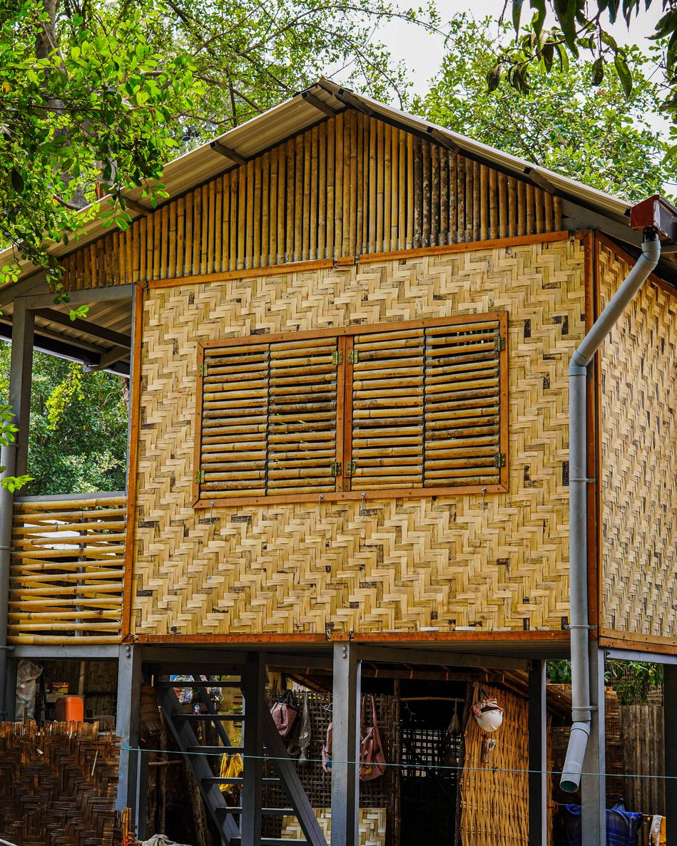 A two-storey mid-term shelter with woven bamboo walls, louvred timber shutters and a metal roof, raised on a steel frame among trees